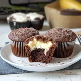 Trzy muffinki czekoladowe, z których jeden został przekrojony, ukazując jasne nadzienie, leżą na białym talerzu.