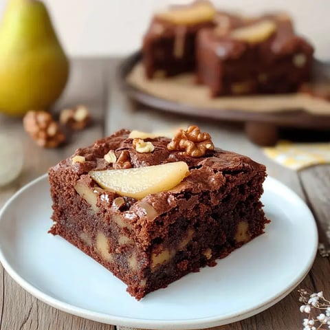 Kawałek czekoladowego brownie z gruszką i orzechami na białym talerzu.